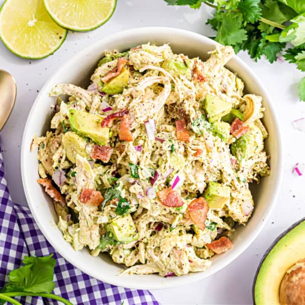 A bowl of chicken salad with avocado, red onion, bacon, and herbs, surrounded by lime slices and cilantro.