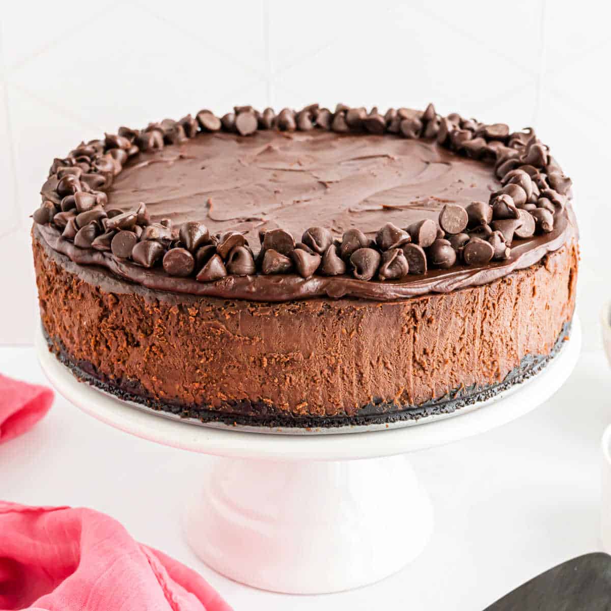 Chocolate cheesecake on a white cake stand, topped with chocolate chips and surrounded by a pink cloth.
