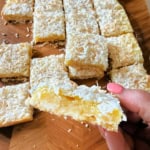 A hand holding a coconut lemon bar; more coconut lemon bars are arranged on a wooden surface in the background.