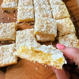 A hand holding a coconut lemon bar; more coconut lemon bars are arranged on a wooden surface in the background.