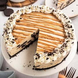 A peanut butter pie with Oreo crust, whipped cream, caramel drizzle, and a slice missing on a cake stand.