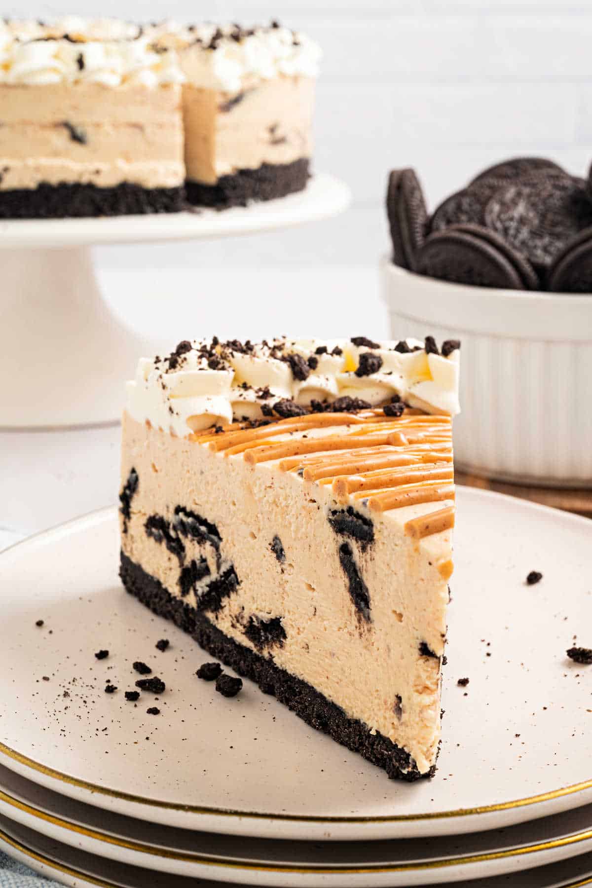 A slice of cookies and cream cheesecake on a plate, with the whole cake and cookies in the background.