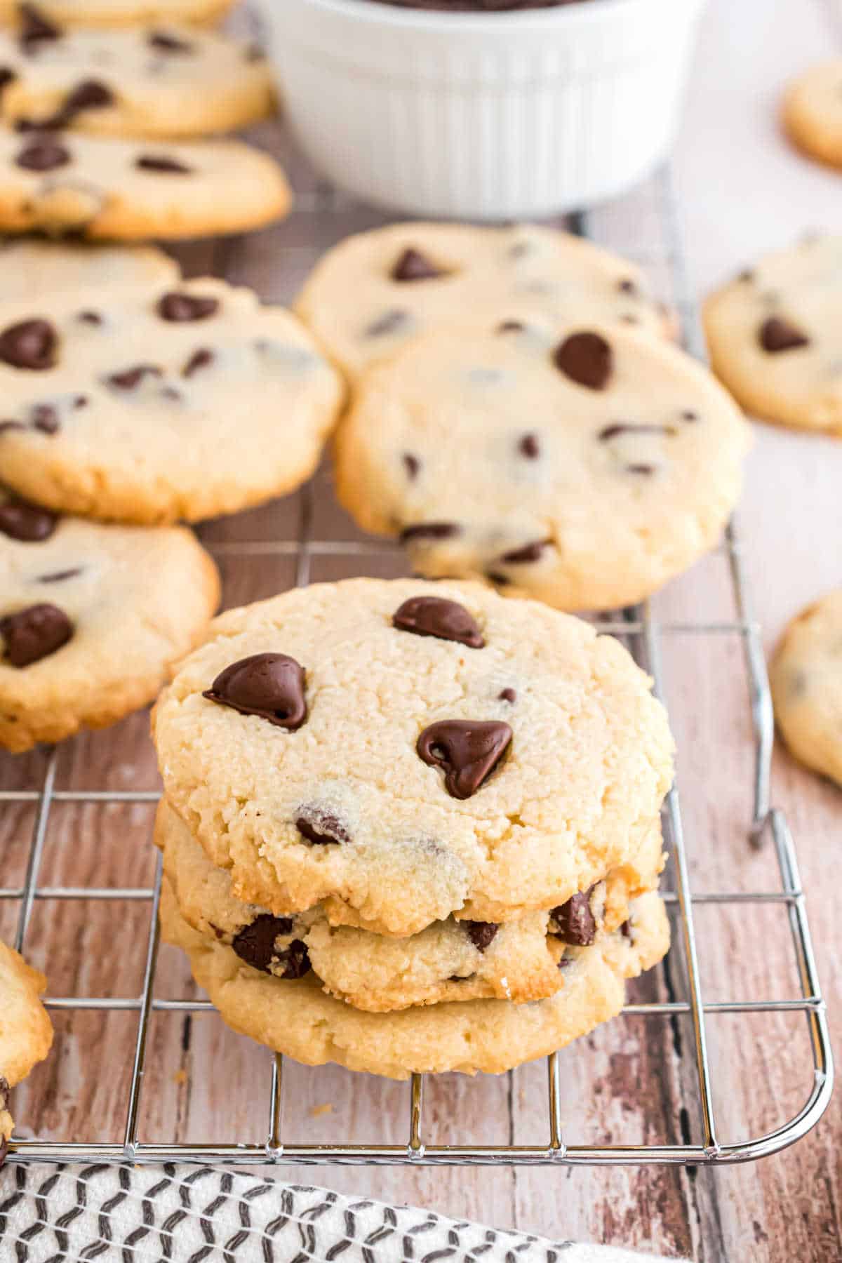 Three chocolate chip cookies stacked on a cooling rack, with more cookies and a bowl in the background.