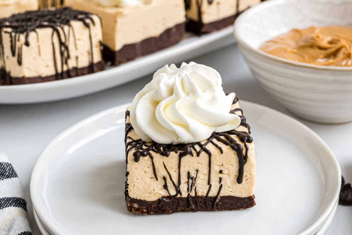 Square peanut butter cheesecake topped with whipped cream and chocolate drizzle on a white plate.