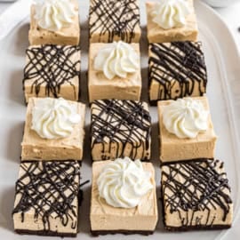 Twelve square dessert bars with a creamy topping, some with chocolate drizzle, arranged neatly on a platter.