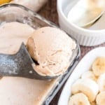 A metal scoop serving banana ice cream from a glass dish, with sliced bananas nearby.