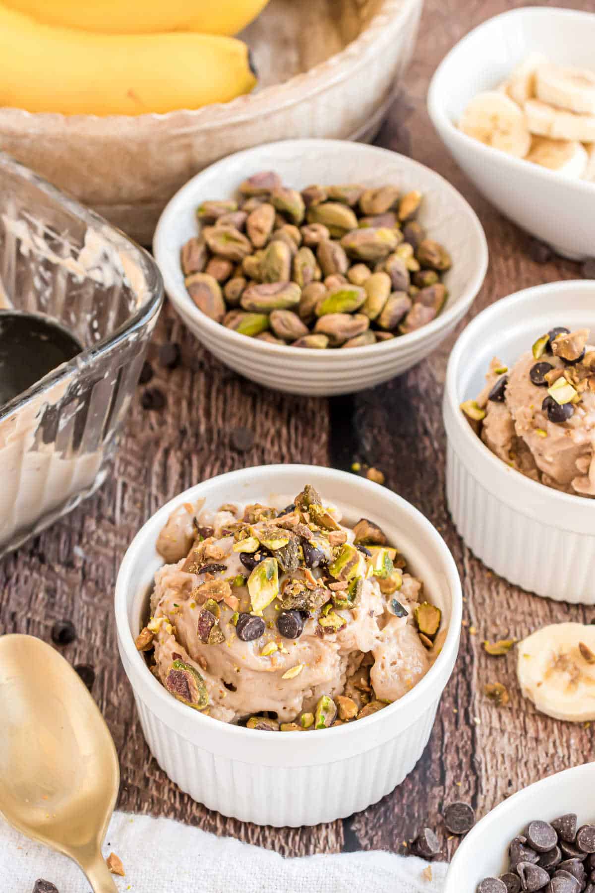 Banana ice cream in a ramekin topped with chocolate chips and pistachios, with bananas and nuts in the background.