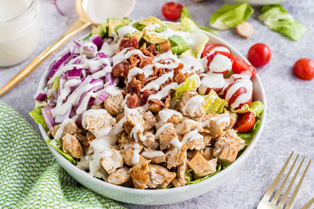 A salad bowl with chicken, bacon, red onions, cherry tomatoes, avocado, and ranch dressing on top.