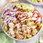 A salad bowl with grilled chicken, bacon, avocado, red onion, tomatoes, lettuce, and ranch dressing.