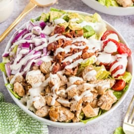 A salad bowl with grilled chicken, bacon, avocado, red onion, tomatoes, lettuce, and ranch dressing.