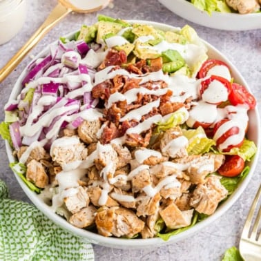 A salad bowl with grilled chicken, bacon, avocado, red onion, tomatoes, lettuce, and ranch dressing.