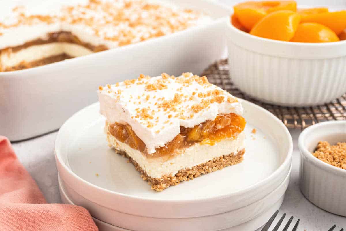 A slice of creamy peach dessert with layers and crumb topping on a plate, with peaches and crumbs in bowls nearby.