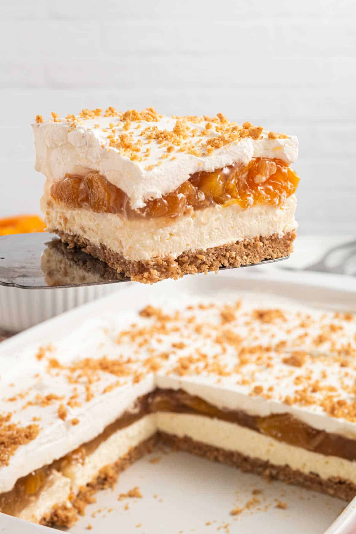 A slice of layered peach dessert with whipped topping and crumb crust being lifted from a pan.