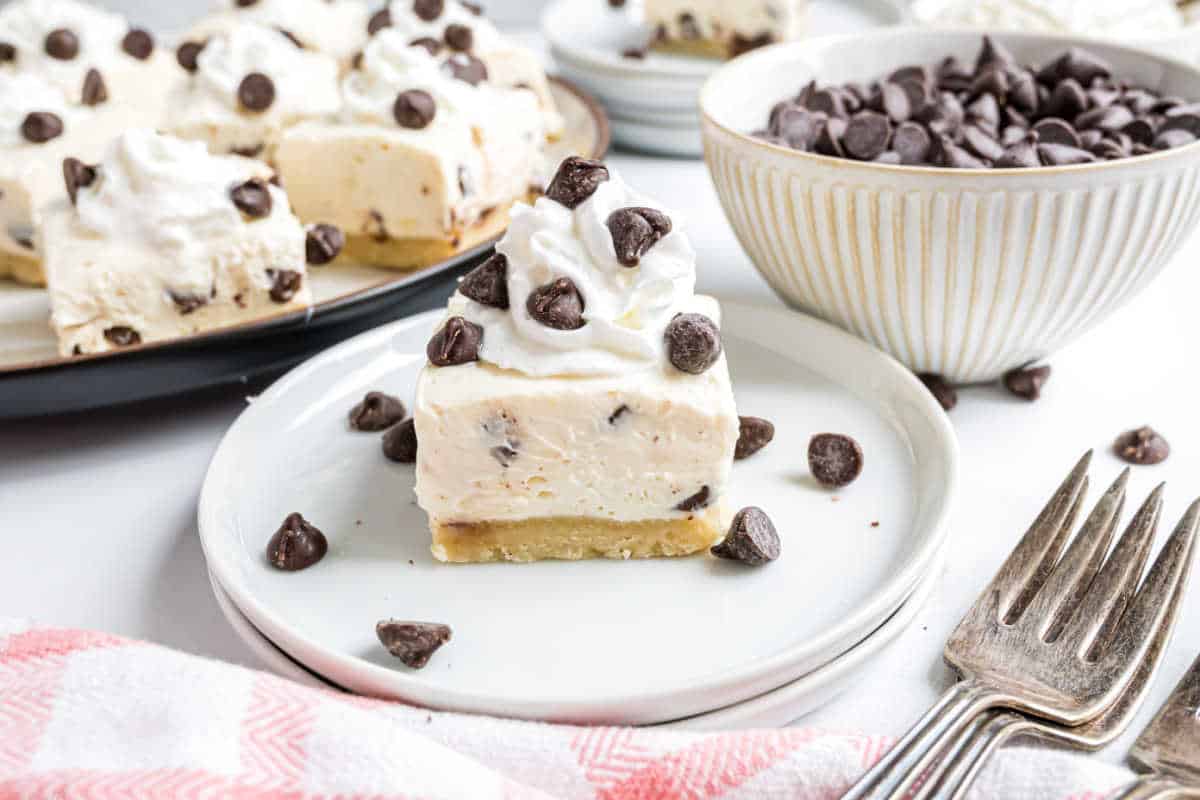 A cheesecake square with whipped cream and chocolate chips on a plate, next to a bowl of chocolate chips.
