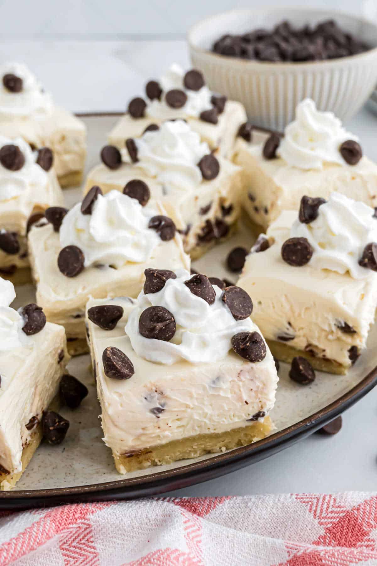 Squares of cheesecake topped with whipped cream and chocolate chips on a plate, with a bowl of chocolate chips nearby.