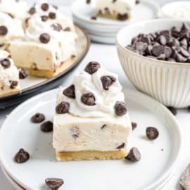 A cheesecake bar with whipped cream and chocolate chips on a plate, surrounded by more chocolate chips.