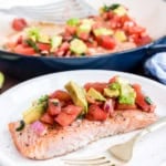 Baked salmon fillet topped with avocado and tomato salsa on a white plate with a fork.