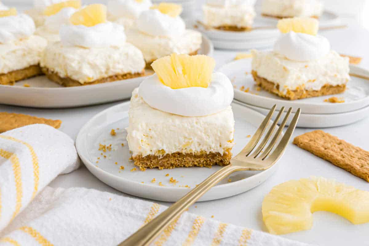 Square pineapple cheesecake slice with whipped cream and pineapple on a plate, fork beside, and more slices in background.
