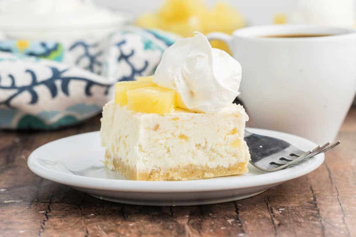 A slice of pineapple cheesecake topped with whipped cream and pineapple chunks on a plate with a fork.