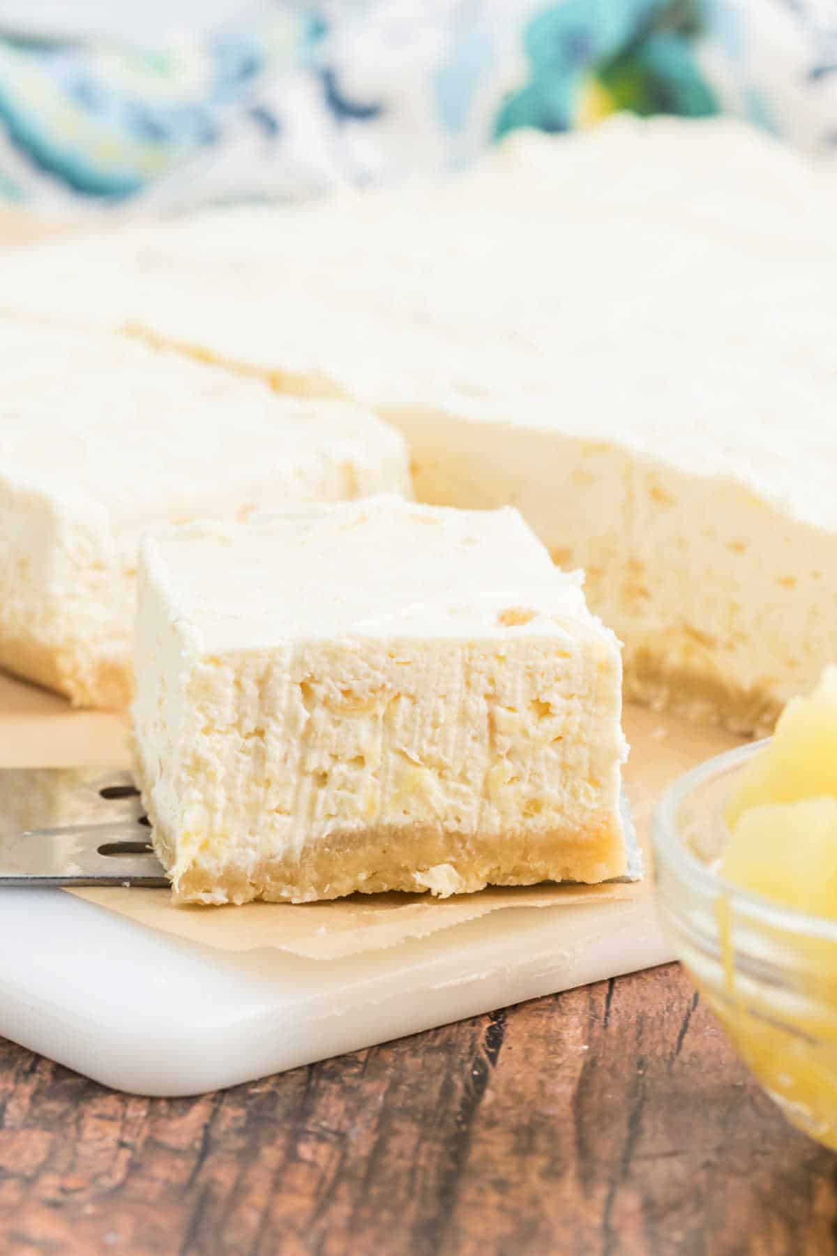 A creamy, square pineapple dessert bar on a spatula, with more bars and a bowl of pineapple in the background.