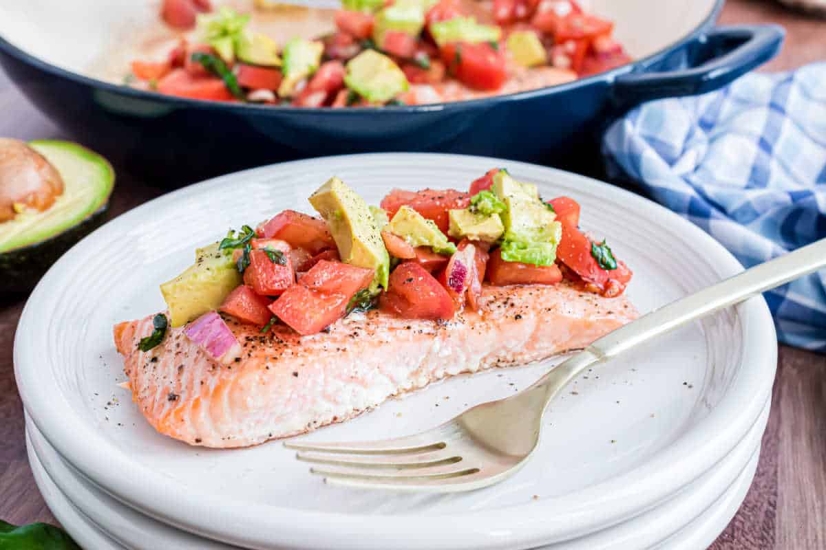 A cooked salmon fillet topped with diced avocado, tomato, and herbs on a white plate with a fork.