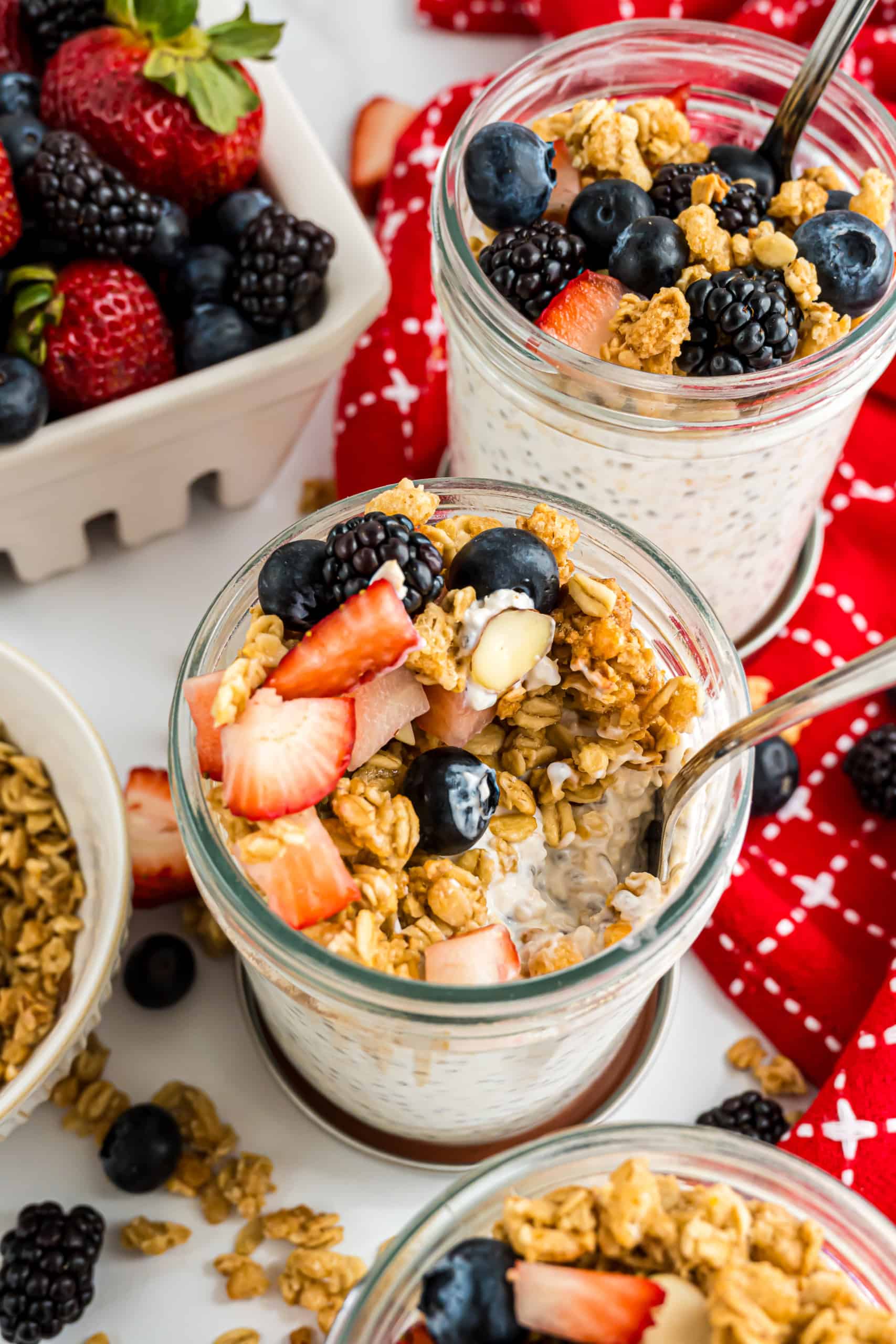 Jars of overnight oats topped with berries and granola, surrounded by fresh fruit and a red napkin.