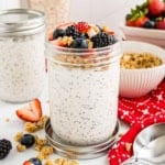 A jar of overnight oats topped with berries and granola, surrounded by more berries and a red napkin.
