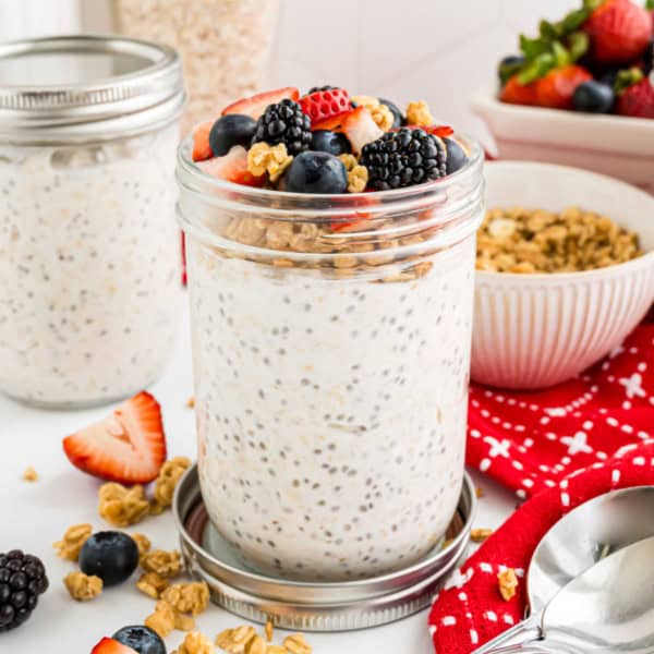 A jar of overnight oats topped with berries and granola, surrounded by more berries and a red napkin.