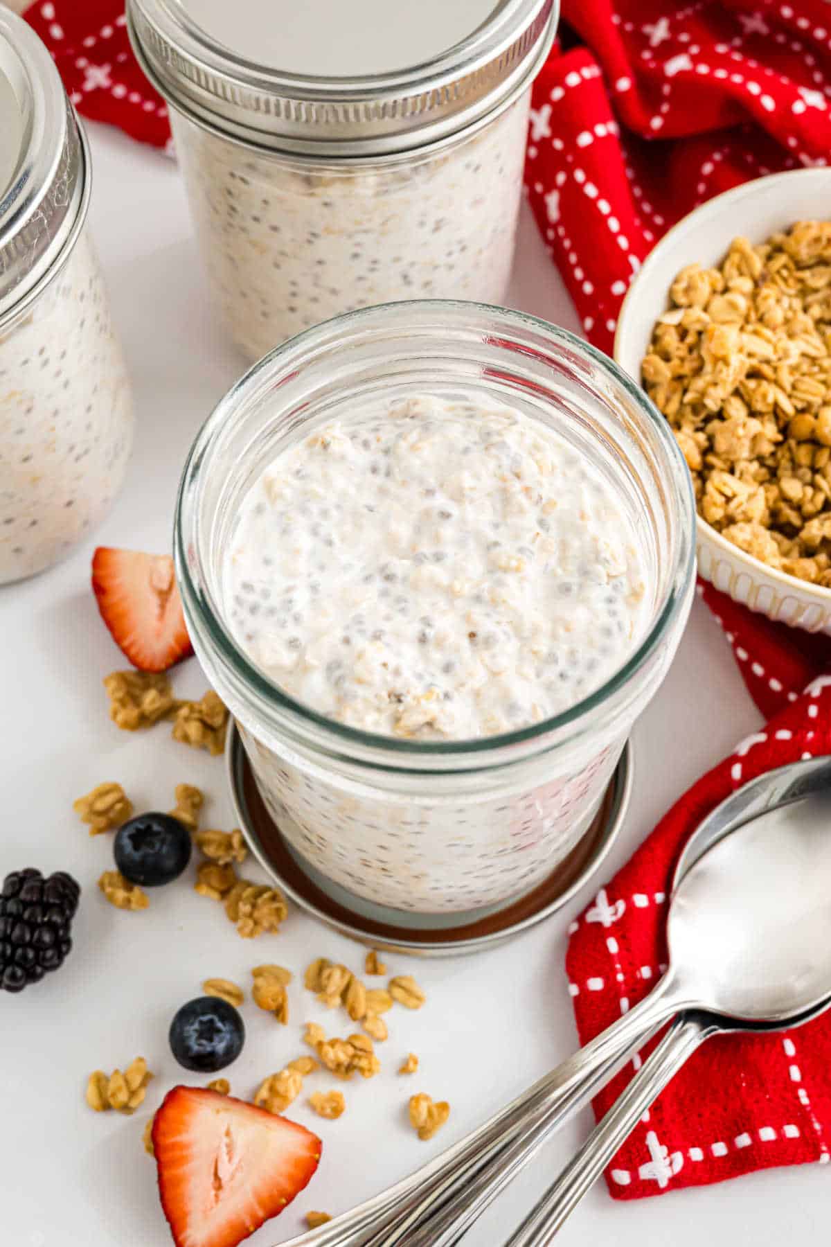 A jar of overnight oats with chia seeds, surrounded by granola, berries, and spoons on a white table.