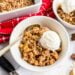 Bowl of apple crisp topped with a scoop of vanilla ice cream, with a spoon and red napkin nearby.
