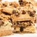 Close-up of crumbly oatmeal raisin cookie cheesecake bars with raisins, stacked on a white surface.